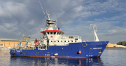 Shows a ship painted blue and white at the harbor entrance. On the side of the ship, you can see the Red Dragon of Wales.