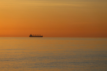 A summer sunrise over a calm Baltic Sea with the silhouette of a ship on the horizon colours the water and the sky deeply red.