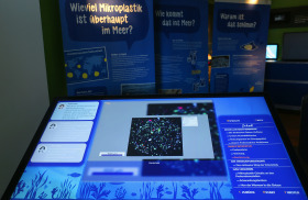 Foto mit Exponaten der Wanderausstellung „Bunt, klein, überall. Mikroplastik – Vom Fluss ins Meer“: ein Multitouch-Tisch und Poster