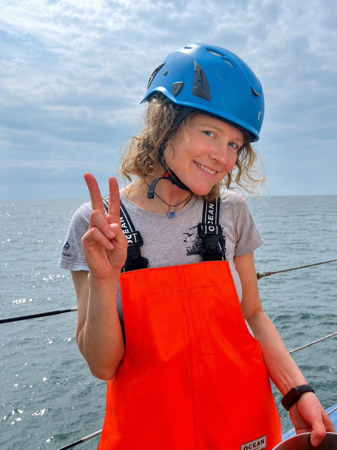 Portrait von Mayya. Sie trägt einen Blauen Helm und eine organge Latzhose. Sie steht an der Reling und hinte rihr sieht man das Meer un den bauen Himmel. Sie macht da Peace-Zeichen.