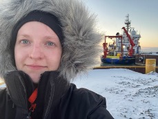 Portrait von Linn. Im Hintergrund liegt Schnee und man sieht ein Foschungschiff am Pier angelegt. Sie trägt einen dicken Manel mit Fellkaputze die das ganze Gesicht einrahmt.